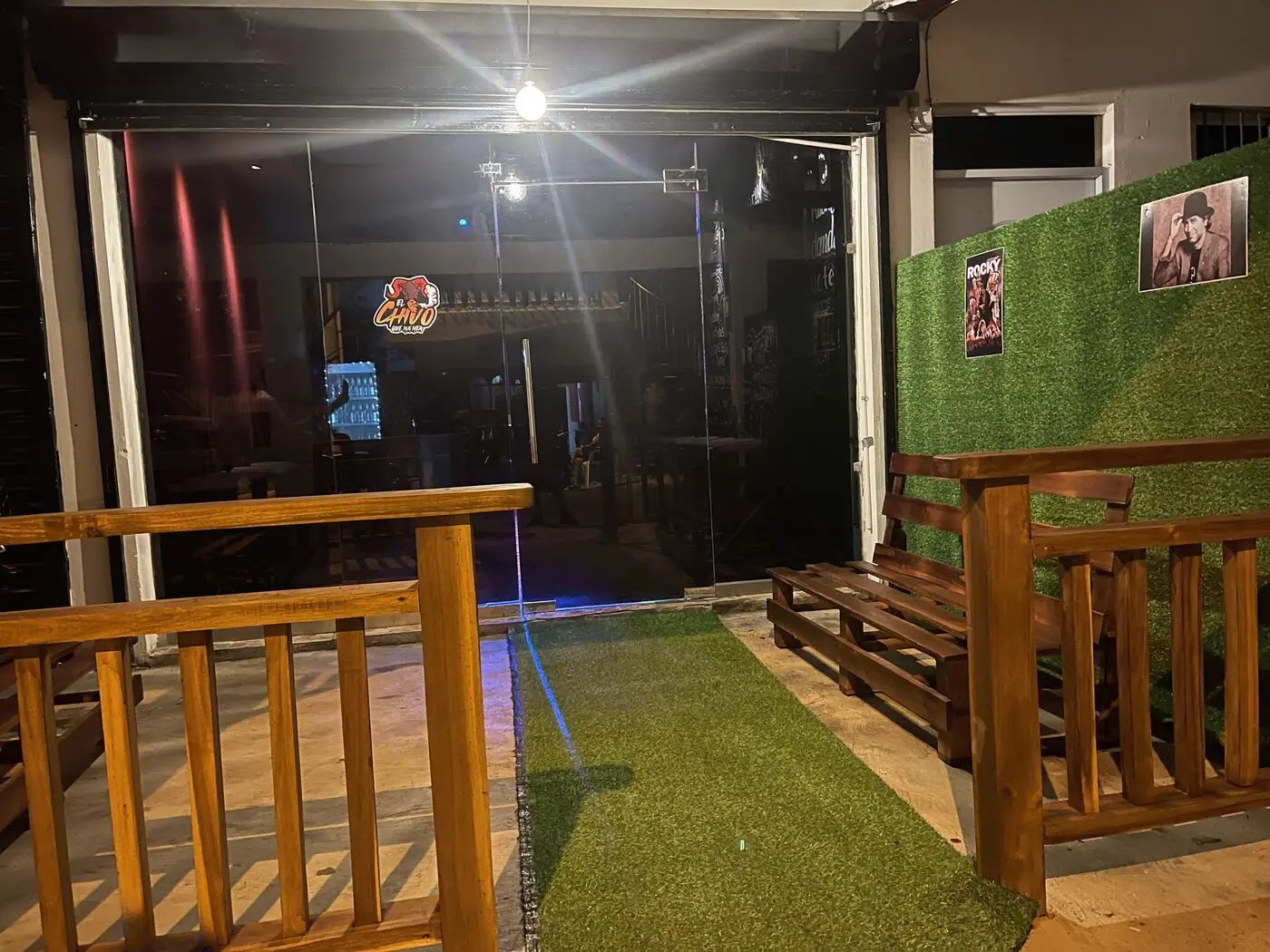 La entrada del bar de noche: grama, bancos de madera y el logo del Chivo en la vidriera