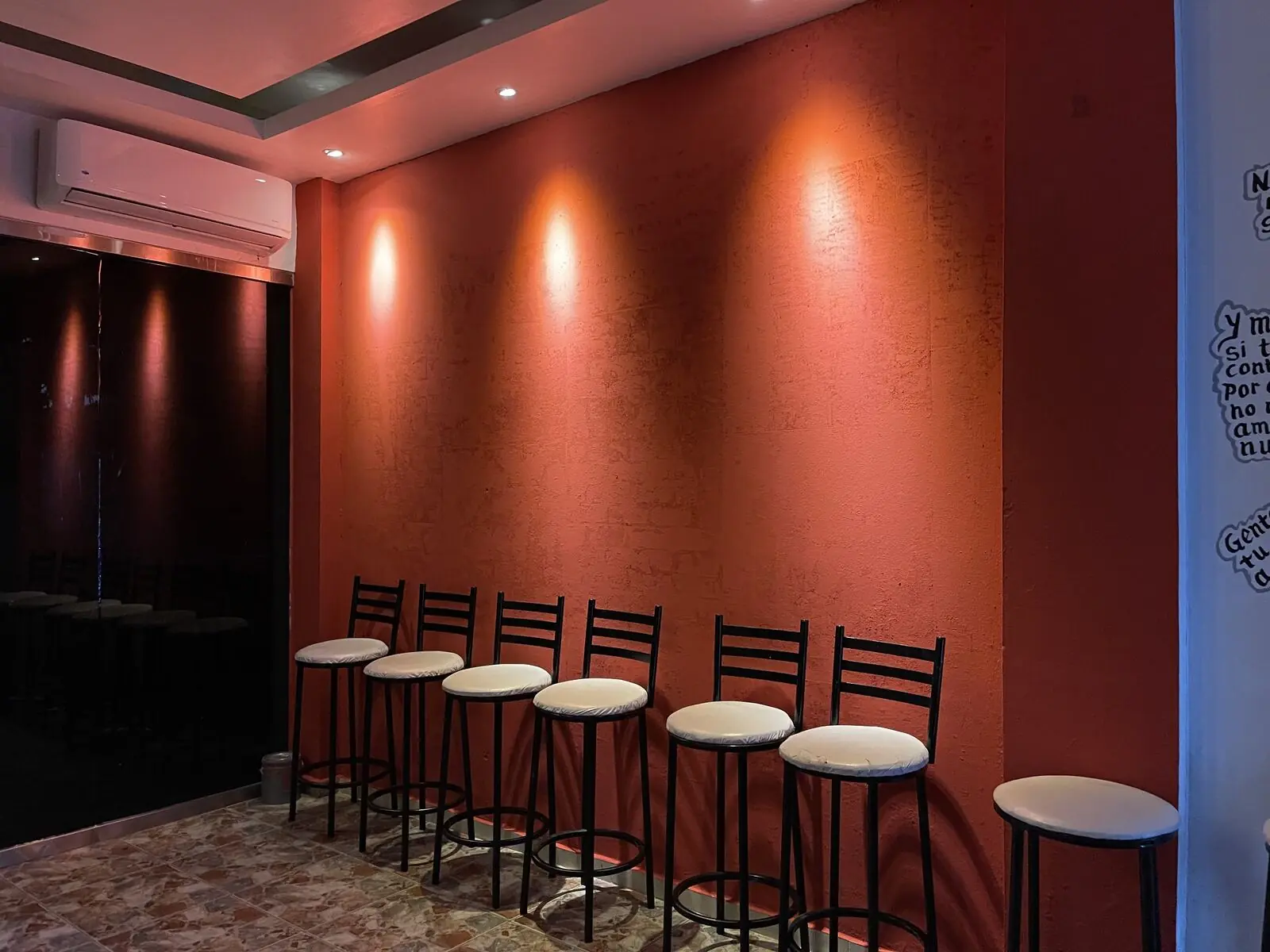 El salón completo del bar: seis banquetas altas contra la pared terracota, con tres luces cálidas encima